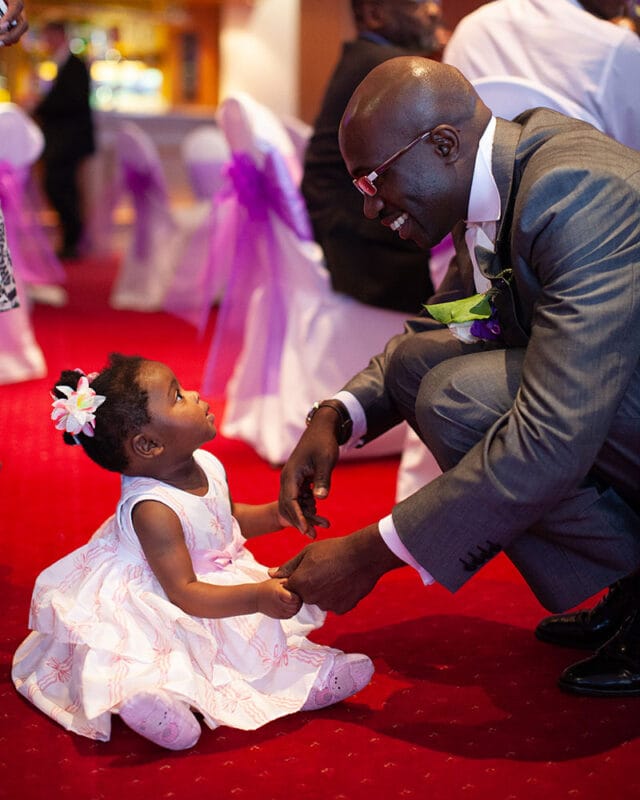 Groom gently holding a young bridesmaid’s hands as she looks up at him with trust and affection