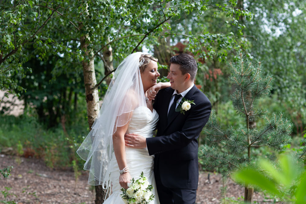 Sam and Tim embrace beside young trees, full of smiles.