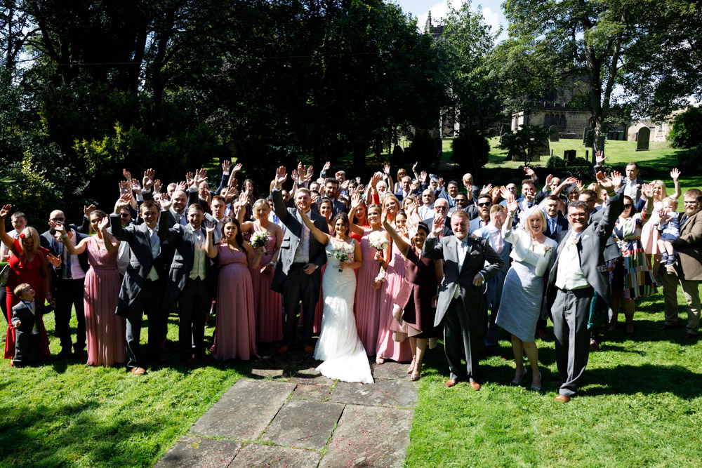 Wide photo of guests waving to the couple in the shade and sunshine of the church gardens.
