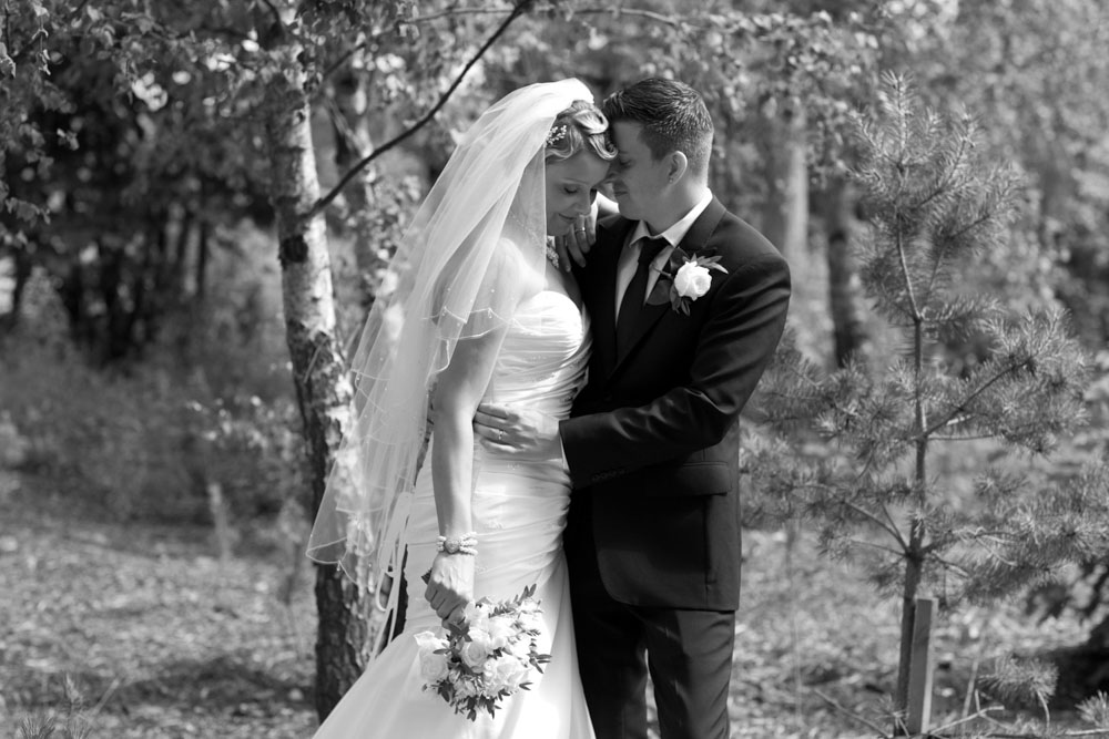 Sam and Tim stood beside young woodland trees in black and white, soft expressions of love.