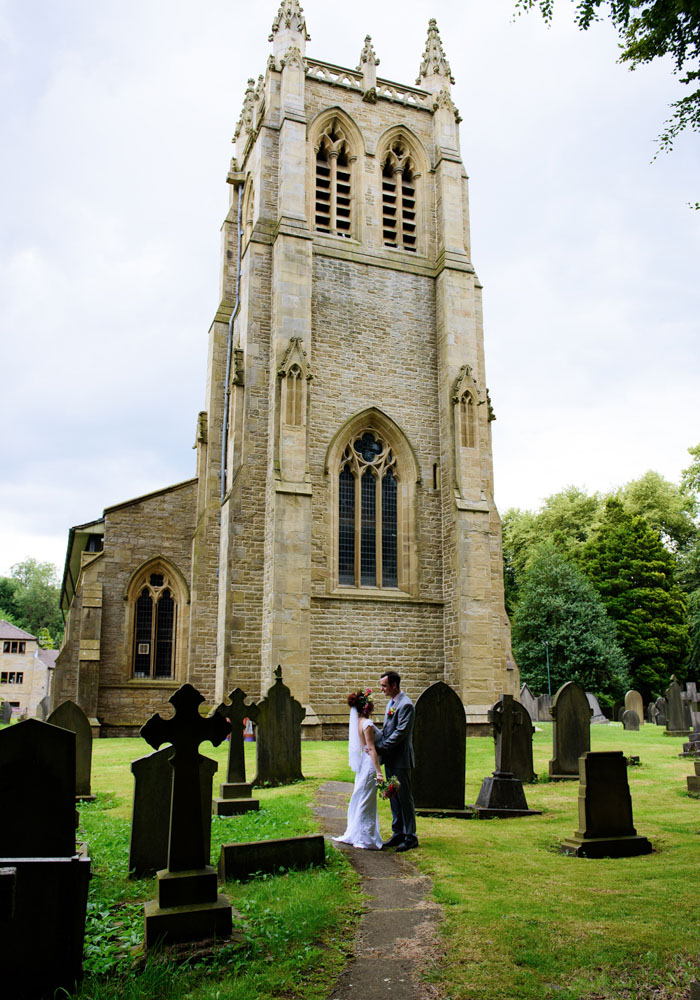 Bride and groom face each other in soft light and shadow, church in the background at Armitage Bridge.