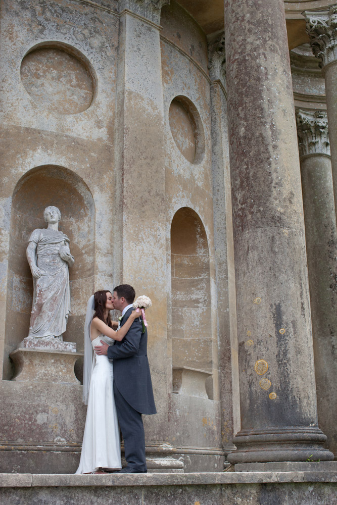 Bride and groom kiss beside a garden statue, bouquet placed playfully on the sculpture’s shoulder