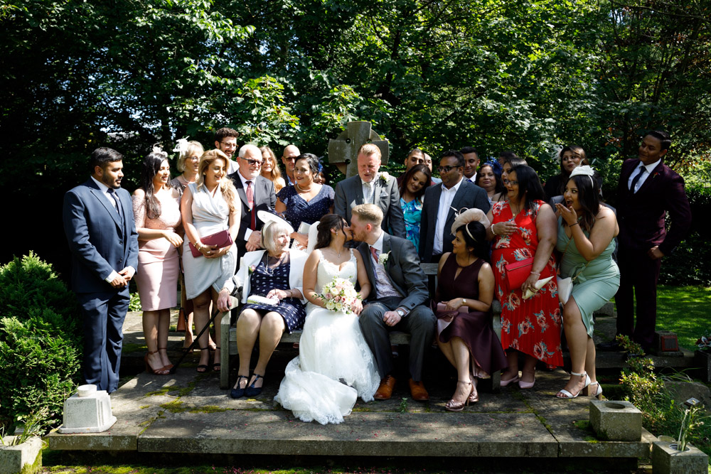 Kyra and Matt kiss during a family group portrait, with relatives smiling and reacting in the background.
