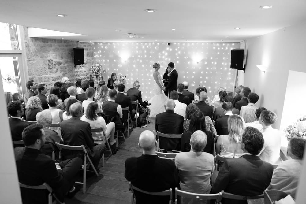Black and white view from the back — couple framed by fairy lights and surrounded by guests.
