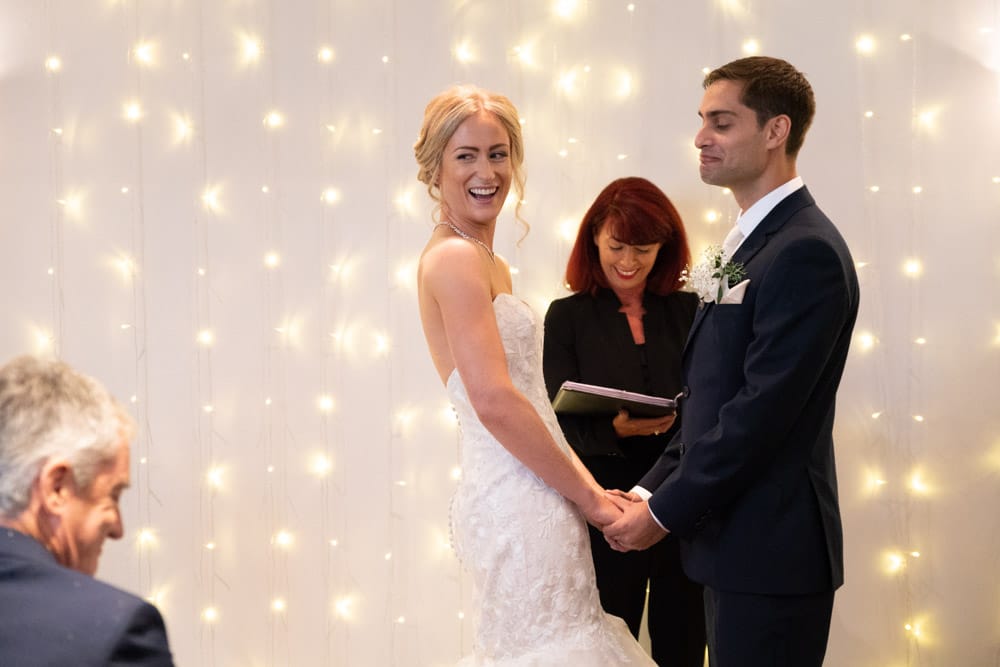 Bride glances back as groom pulls a playful face during Chilli Barn wedding ceremony.