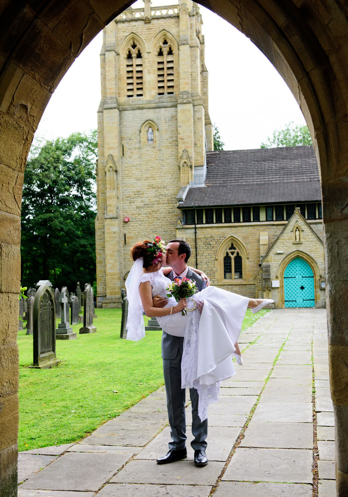 Groom lifts the bride playfully beneath an arch, church behind, bride points her ballet-style shoes.