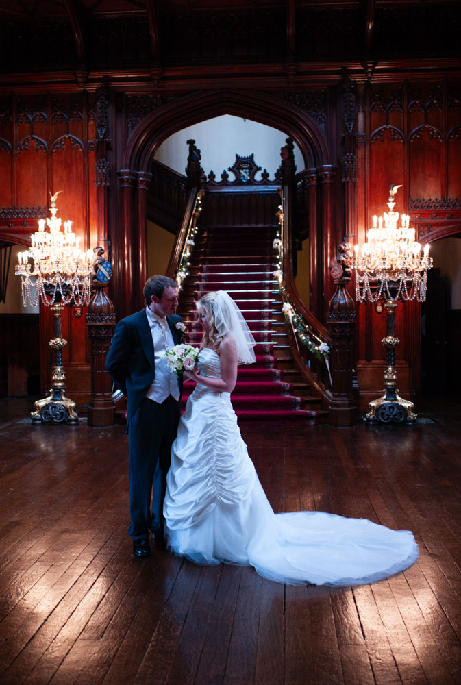 Bride and groom on the staircase at Allerton Castle, lit by lamps and chandelier glow.