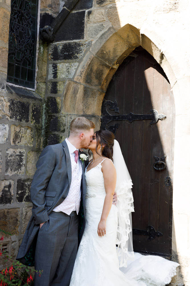 Kyra and Matt kiss by an old wooden side gate beside the stone walls of St Oswald’s Church.
