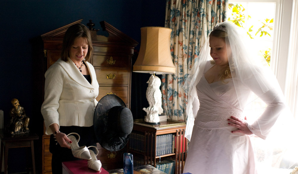 Bride's friend smiles as she gently touches the wedding shoes; the bride watches with hands on hips — a quiet, documentary-style moment full of warmth and anticipation.
