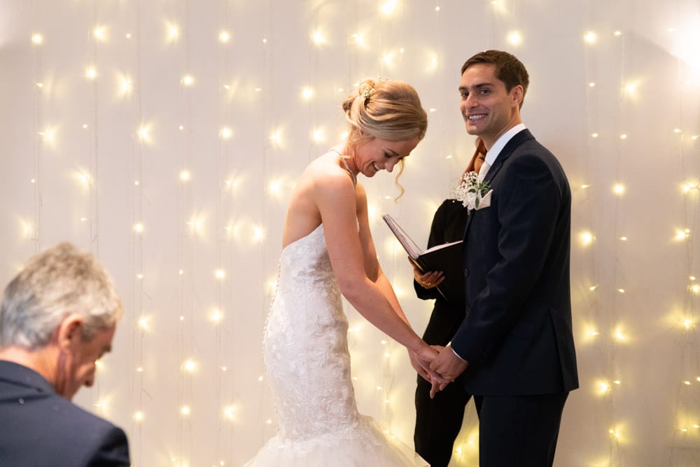 Bride laughing as the groom smiles toward the guests during their Chilli Barn wedding vows.
