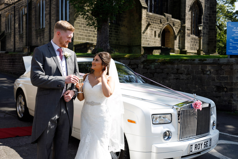 Kyra and Matt clink champagne glasses beside their wedding car, the church visible behind them.