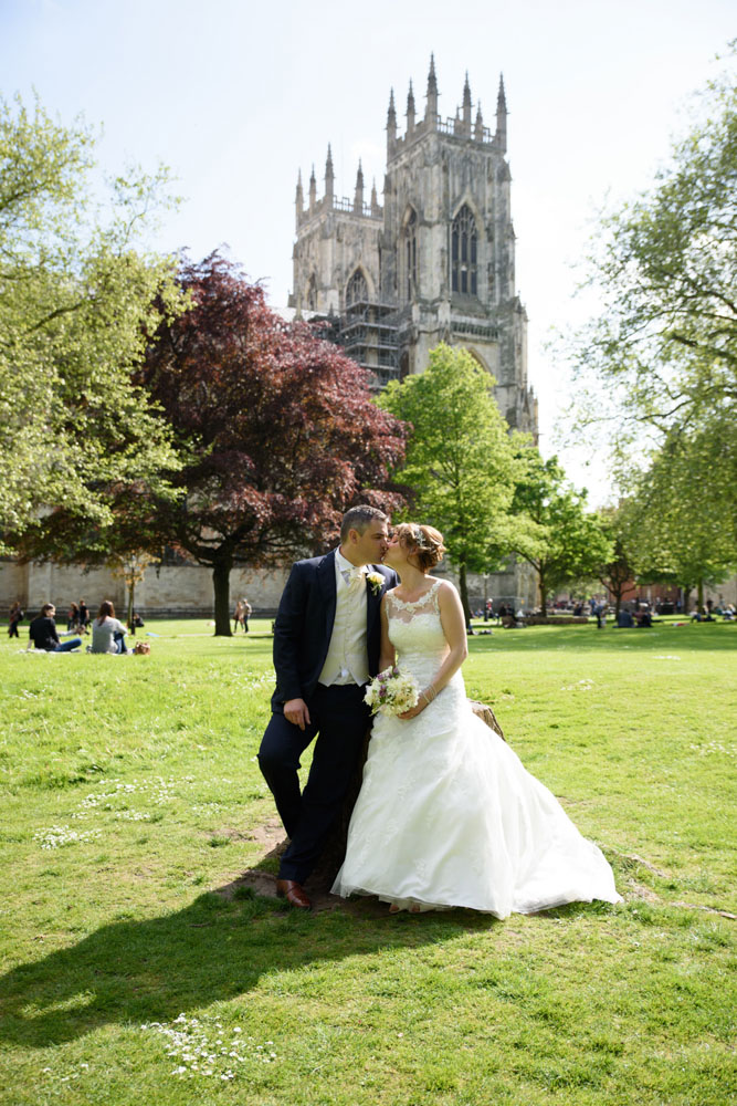 Couple kiss while seated on a wooden stump with trees and York Minster in the background.