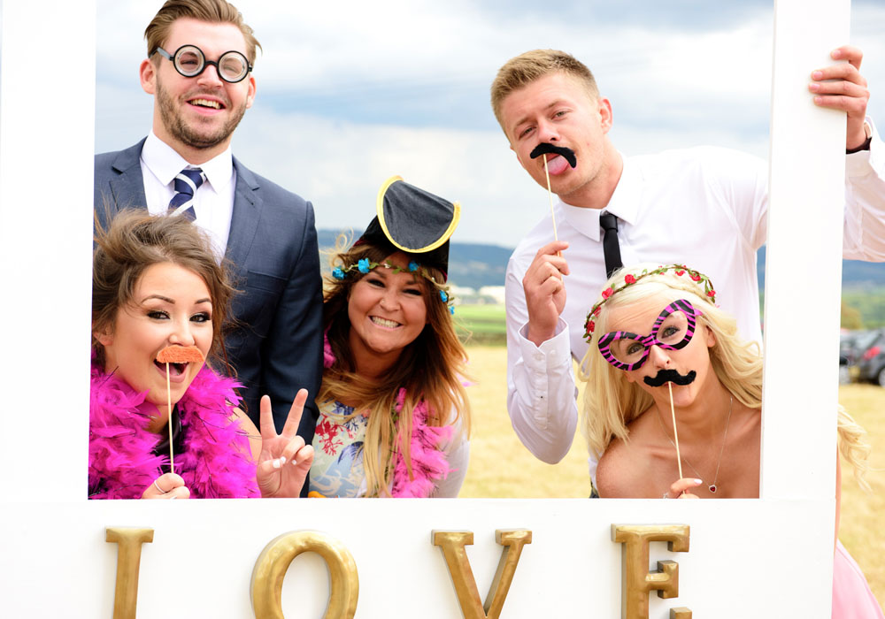 Group of five friends posing in the ‘love’ selfie frame with fun glasses, hats, and moustaches.