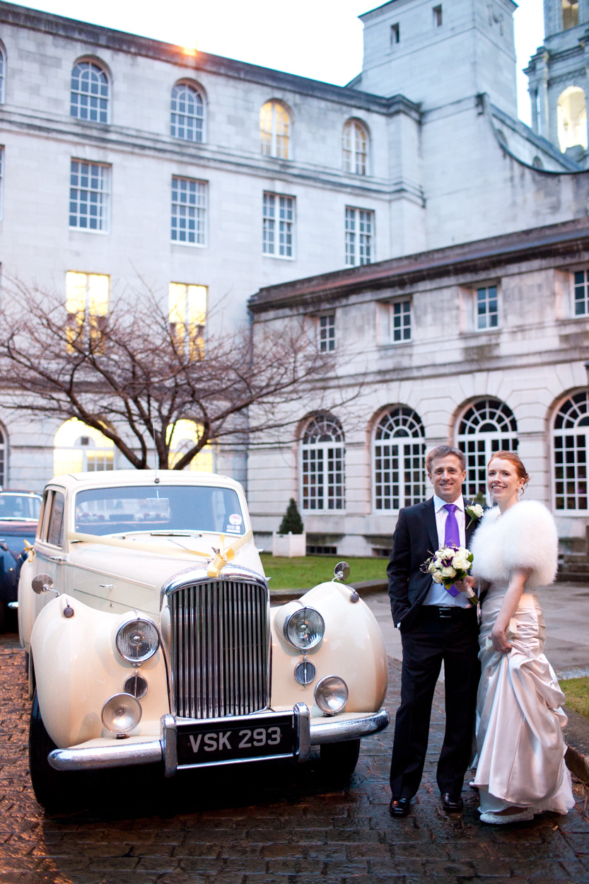 Bride and groom standing together by the wedding car, bare trees and white buildings behind.