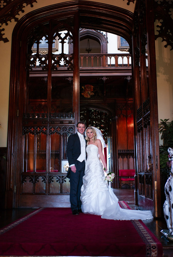The couple stand by tall wooden doors on the dark red rug at the venue entrance