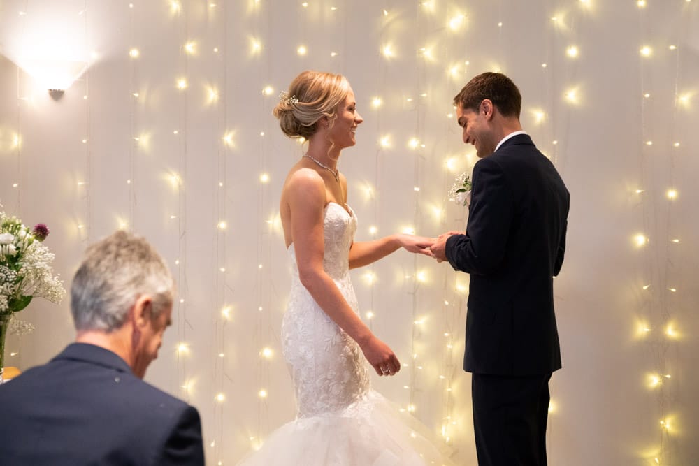 Adam places the wedding ring on Helen’s finger, both smiling with joy.