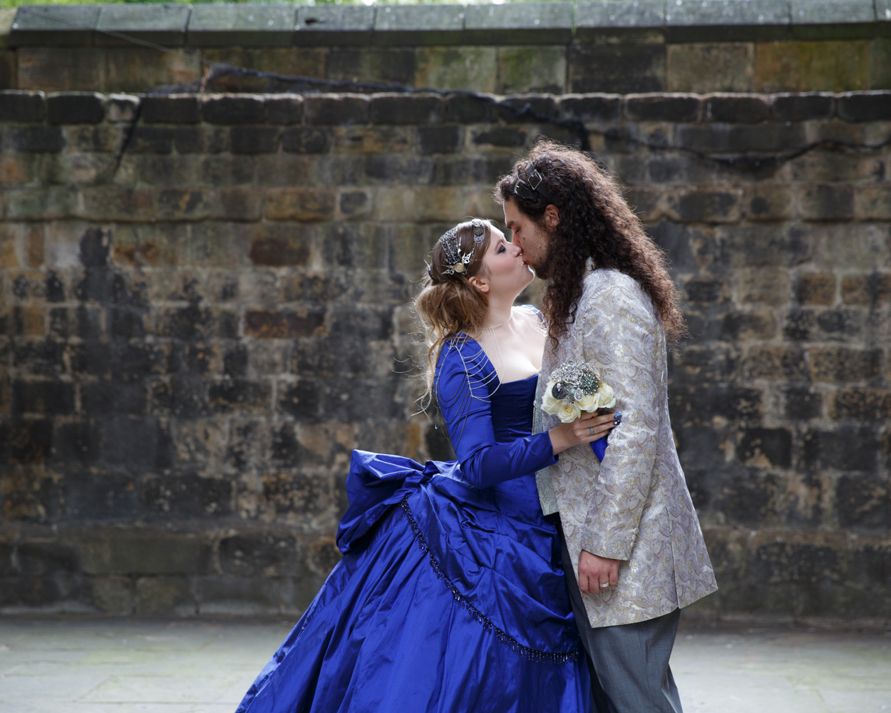 The couple share their first kiss, framed by blue sky and stone walls