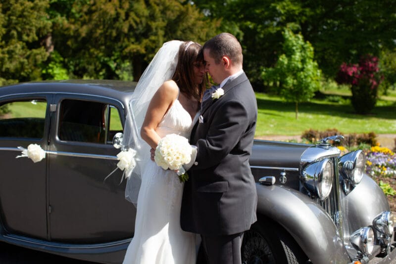 Wedding couple nose to nose in sunshine beside their vintage car, bouquet catching the light