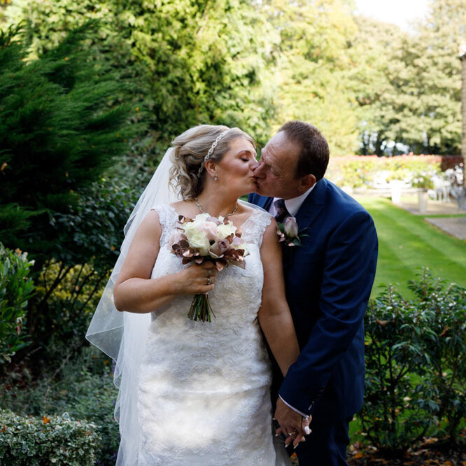 Couple kiss beside the low wall with lawns and trees behind