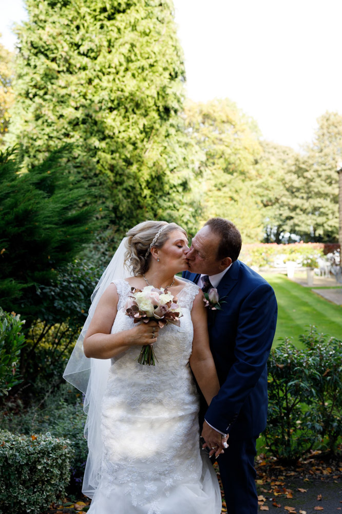 Couple kiss beside the low wall with lawns and trees behind