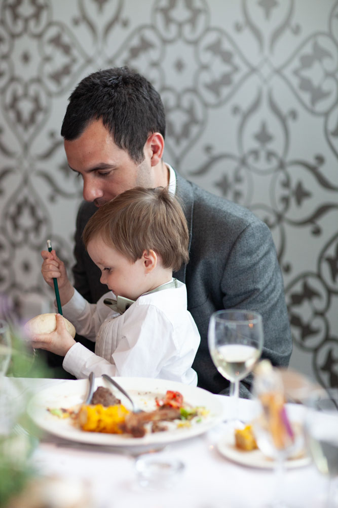 A man helps his little son at the table, the boy trying to cut bread with a pencil.