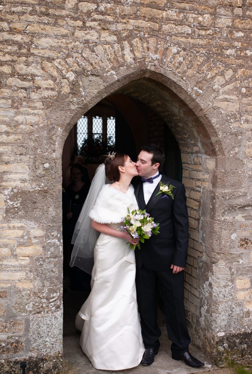 The couple kiss under the stone arch in winter sunlight.