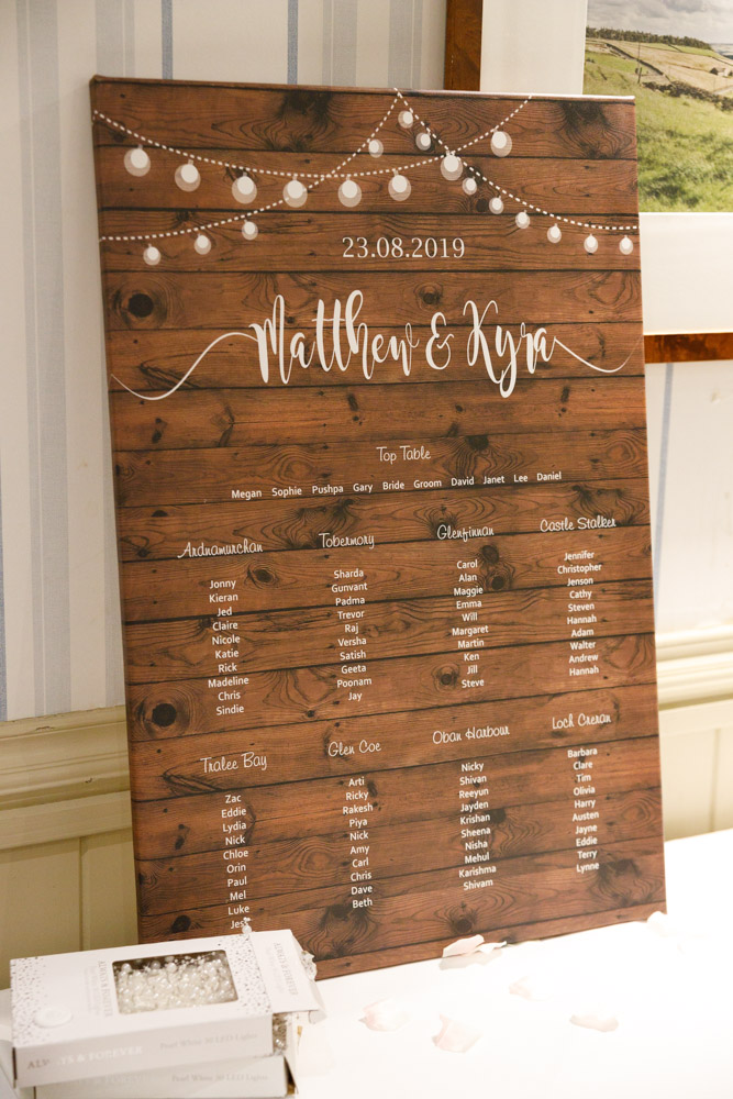 Rustic wood and white table plan reading “23.8.19 Matthew & Kyra” with elegant styling.