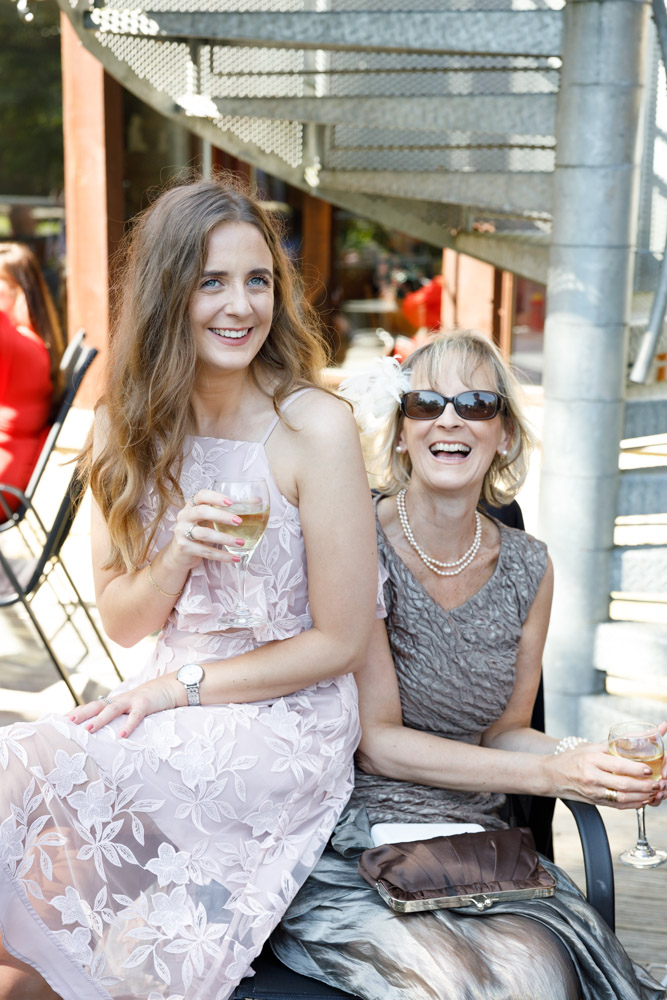 A mother and daughter relax with wine under dappled sunlight outside the wedding venue.