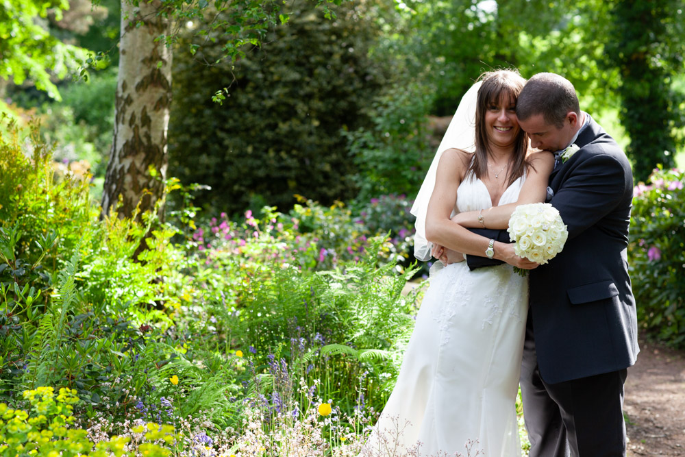 Groom kisses bride’s shoulder in a hidden garden filled with green trees and flowers.