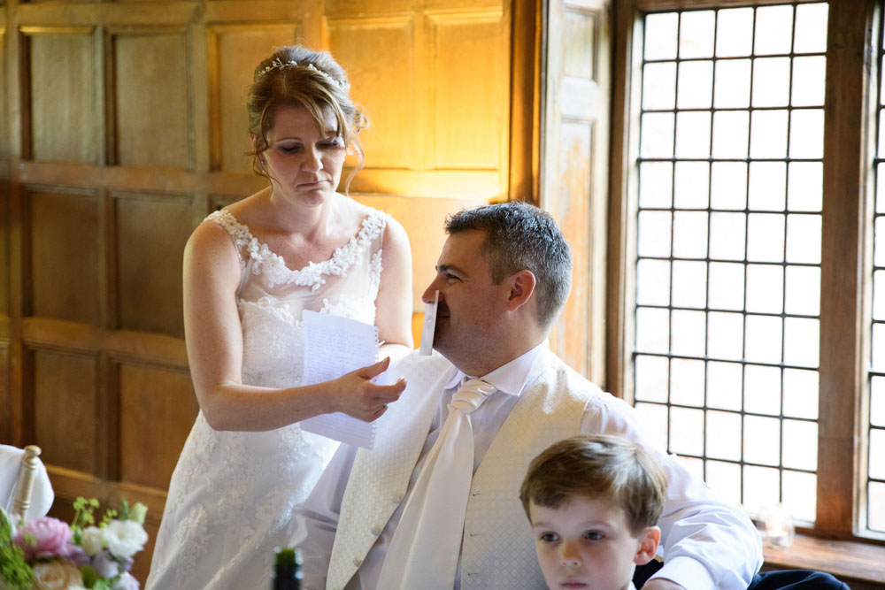 Bride holds a smile card under the groom’s nose, making a joking sad face at him.