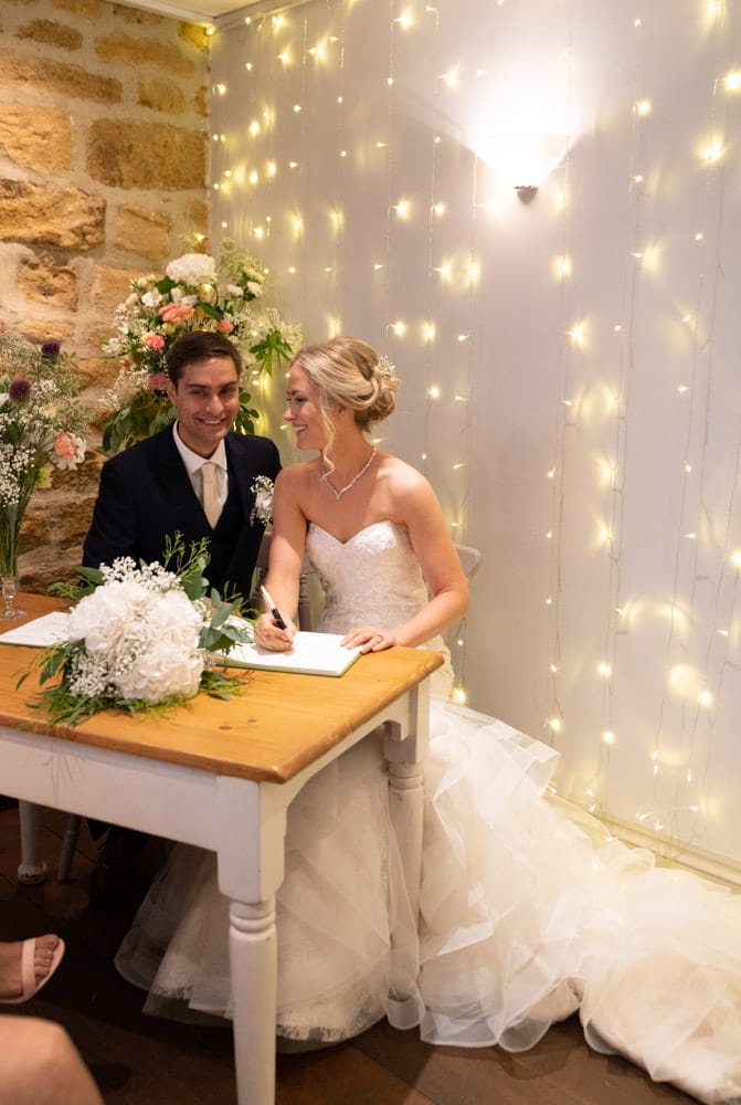Full-length view with fairy lights behind, the couple pose in mock signing, bouquet on the table.