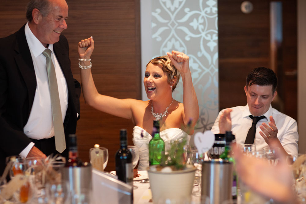 Sam cheers with both arms up as her father begins his speech, joyful energy in the air.