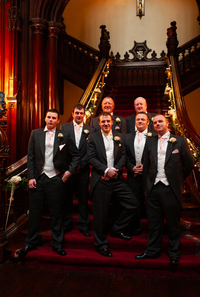 Groomsmen lined up on the grand staircase at Allerton Castle, surrounded by red and gold interiors