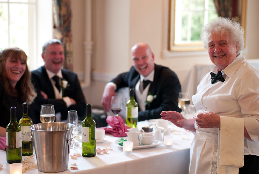A smiling waitress turns toward the camera as guests react with laughter at the table.