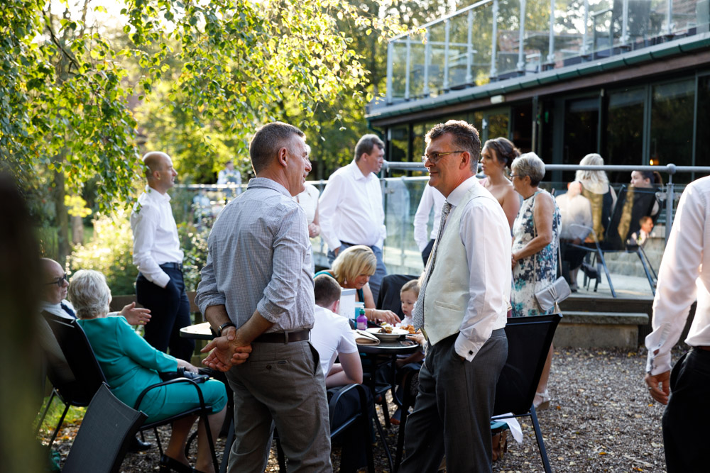 Wedding guests relax and chat in the afternoon sun outside Chevin Country Park Hotel, surrounded by trees and woodland.