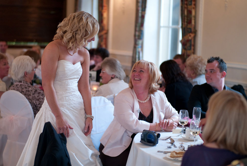 Dani stands in her wedding dress, chatting warmly with a guest seated at their table.