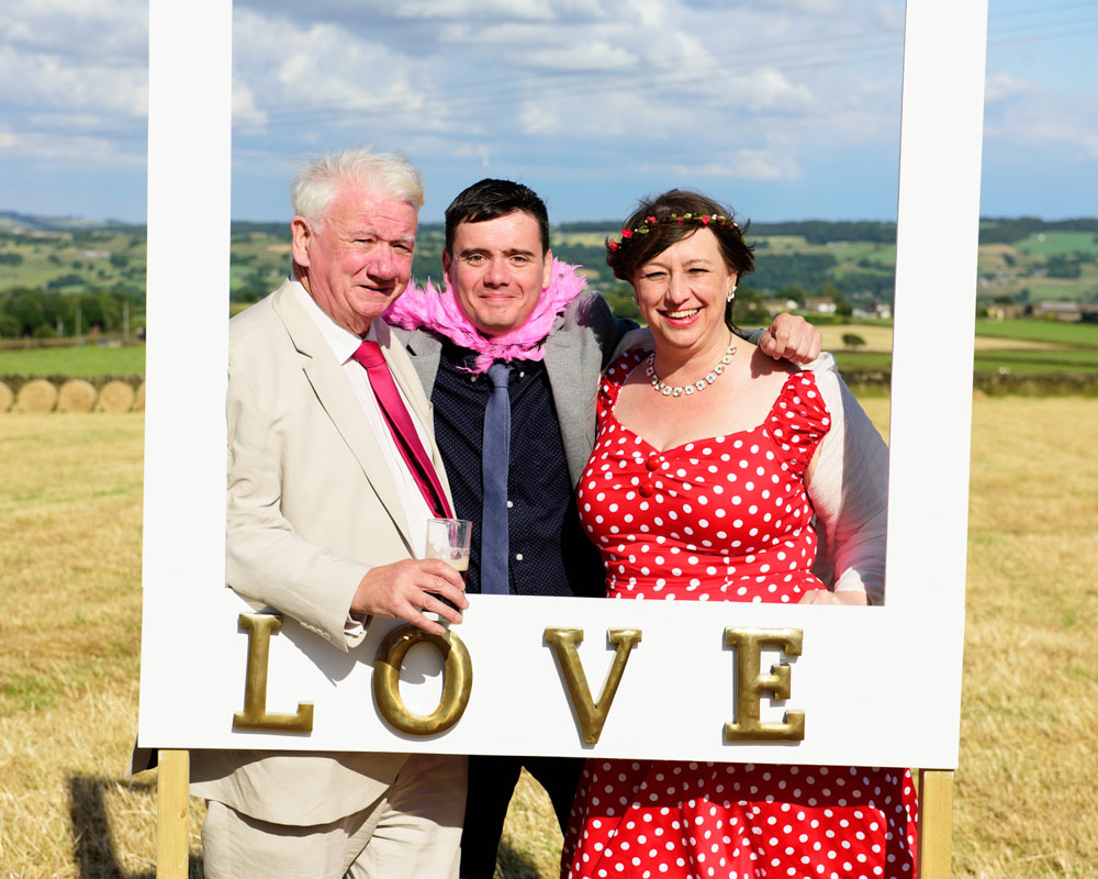 Bride, groom, and their young son pose together in the 'love' selfie frame with countryside behind.