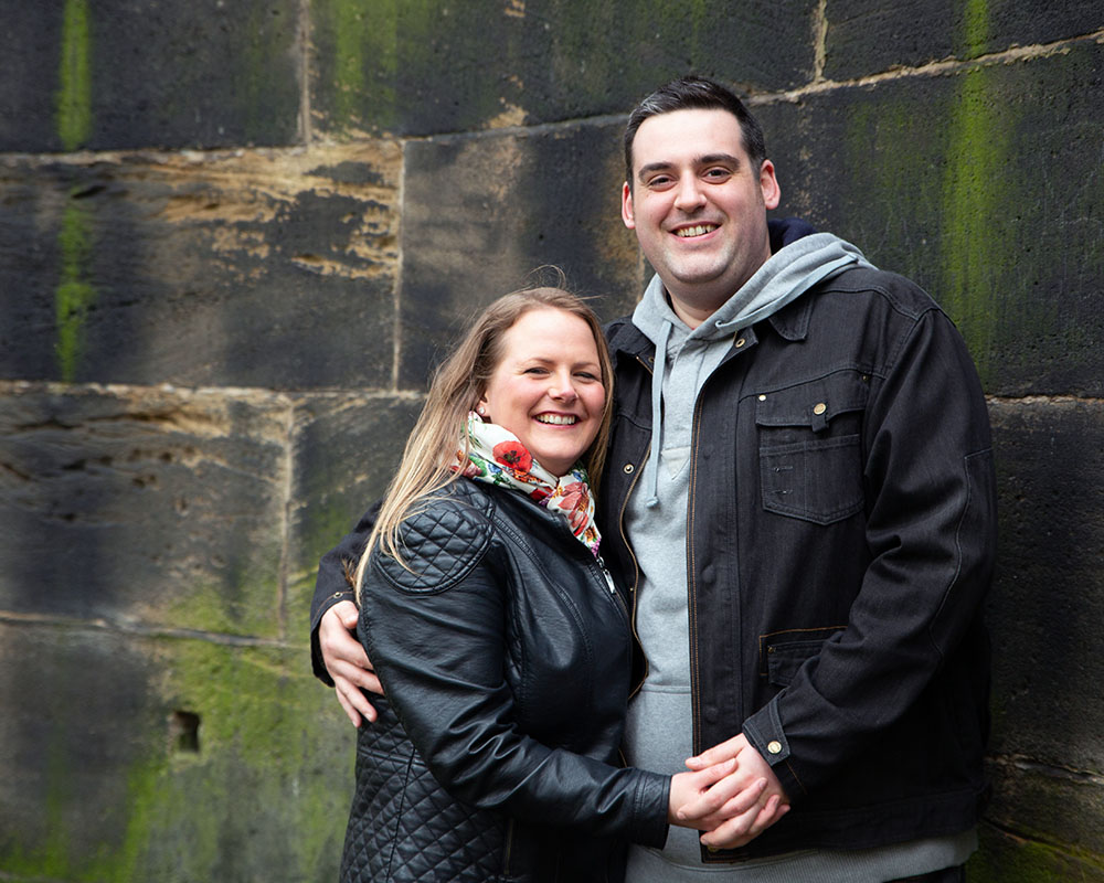 Lisa and Oliver sharing joyful smiles by a moss-covered wall at Kirkstall Abbey.