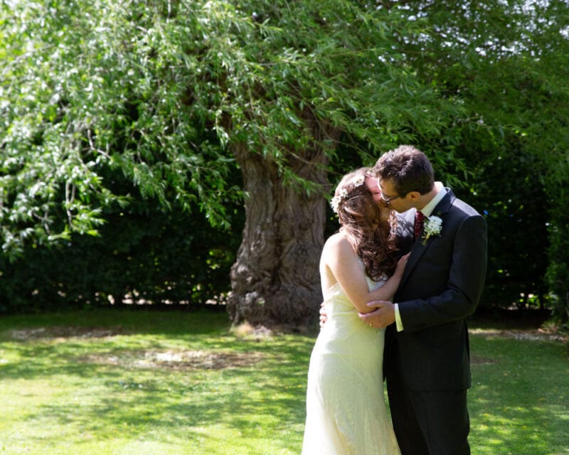 A wedding couple kiss under wind-blown trees, bride’s hair lifting in the breeze