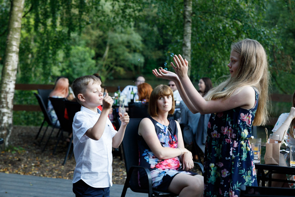 Children play with bubbles while a mum looks on, framed in soft summer shade.