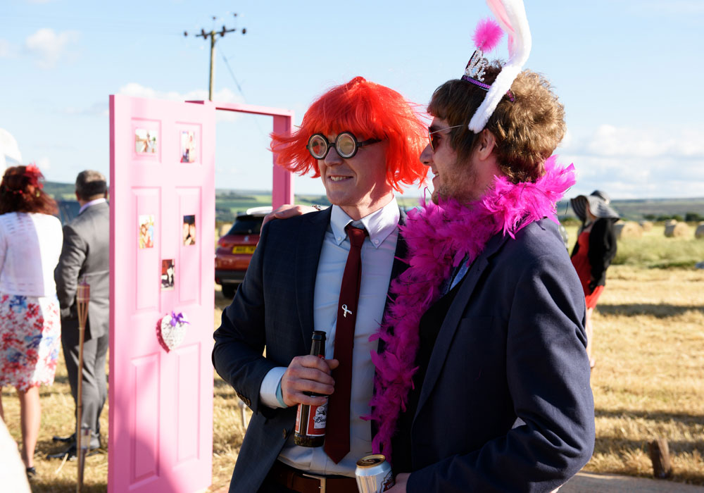 Guest in orange wig, yellow garland, and funny glasses poses at the wedding.
