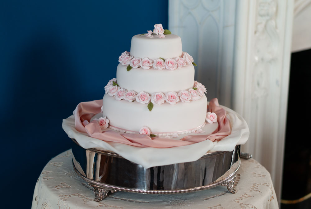 Three-tiered wedding cake with icing roses on silver stand, Allerton Castle wedding breakfast room behind