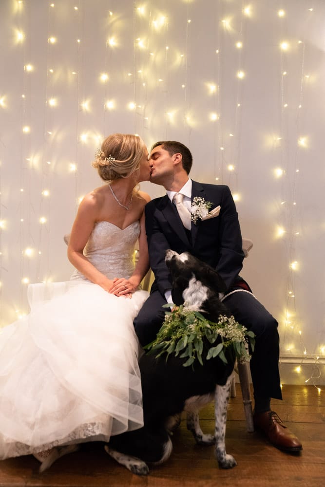 The bride and groom kiss as their dog looks up at them curiously.