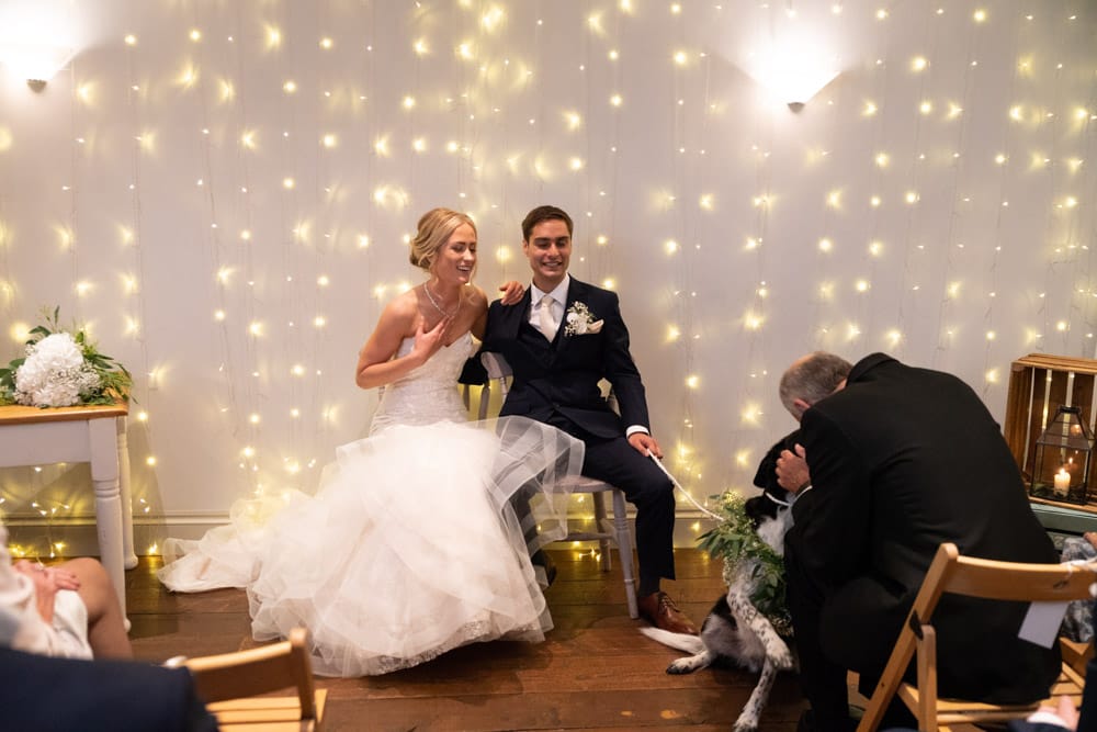 Wide shot of the couple seated beneath fairy lights as their dog greets the groom’s son.