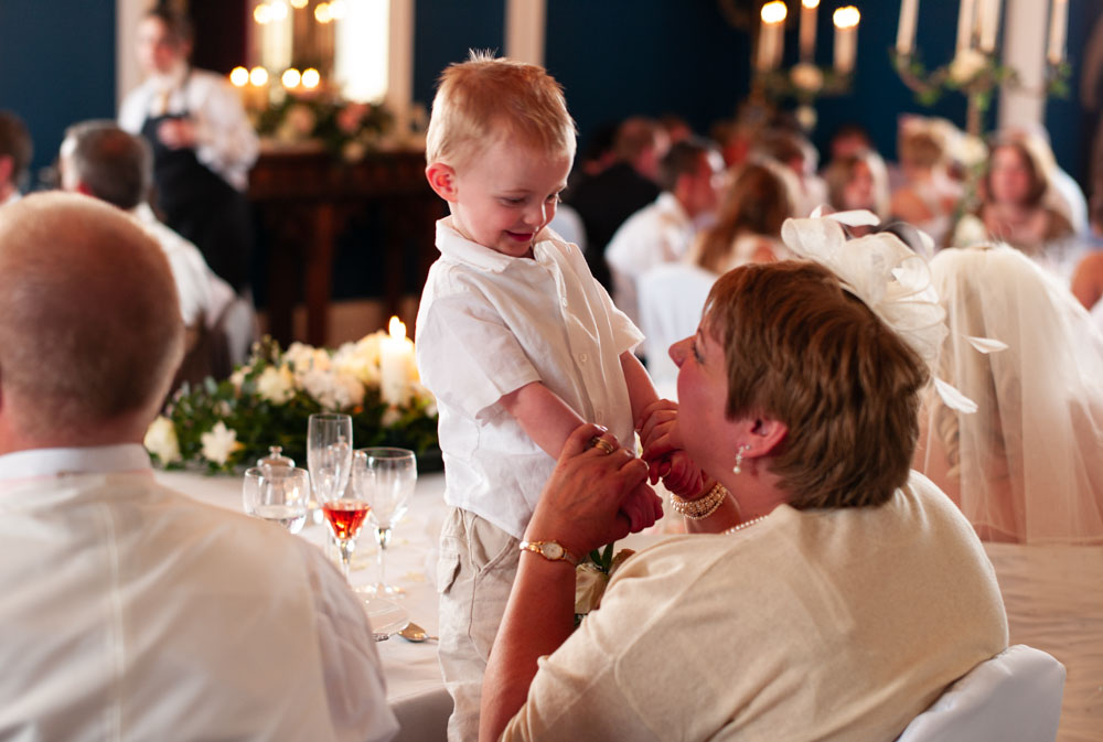Mother of the bride holds her smiling grandson on her lap, standing hand in hand