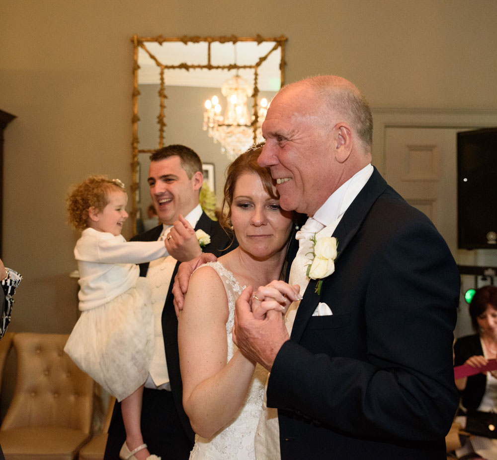 Bride and her father share a dance, with the groom and child watching behind.