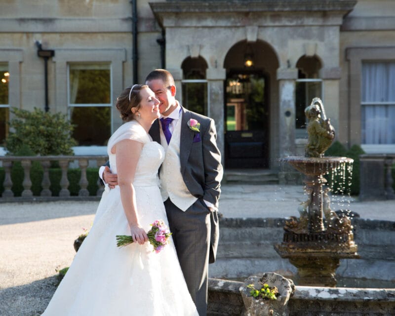 Groom with a shy, gentle smile by a decorative fountain outside a historic stone venue in golden hour light