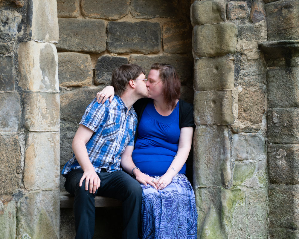 A quiet kiss between Rachel and Robert seated in the stone archway at Kirkstall Abbey.