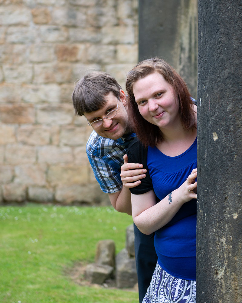 Rachel and Robert peeking out from behind a stone column, sharing playful smiles.