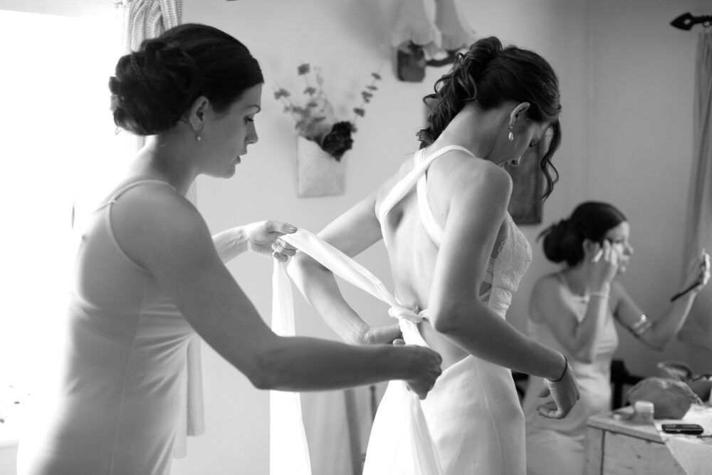 In black and white a bridesmaid helps tie the back of the bride's elegant wedding dress - another bridesmaid doing makeup sat at the table